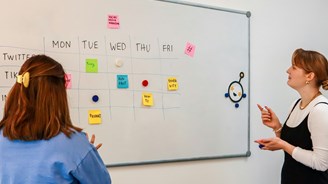 Two women planning social media content for accountants on a whiteboard calendar with sticky notes labeled Twitter, TikTok, Instagram, and various content ideas for each day of the week.